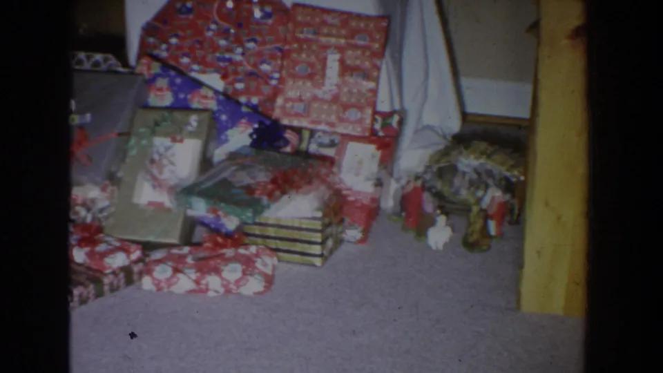 1965 Fallston, Maryland home-movie frame showing wrapped Christmas gifts, bows, carpet, and small seasonal figurines indoors.