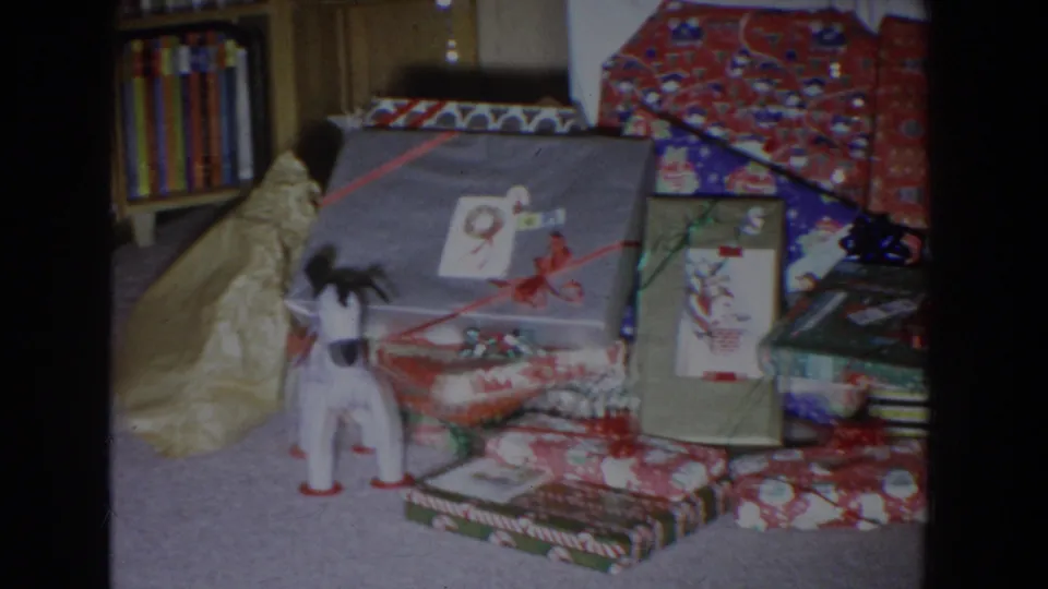 1965 Fallston, Maryland home-movie frame showing wrapped gifts, ribbon, a toy dog, and bookshelves in a private holiday interior.