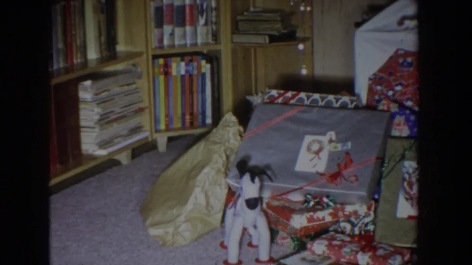 A small white and black toy dog stands in front of wrapped Christmas presents in a 1965 Fallston home interior.