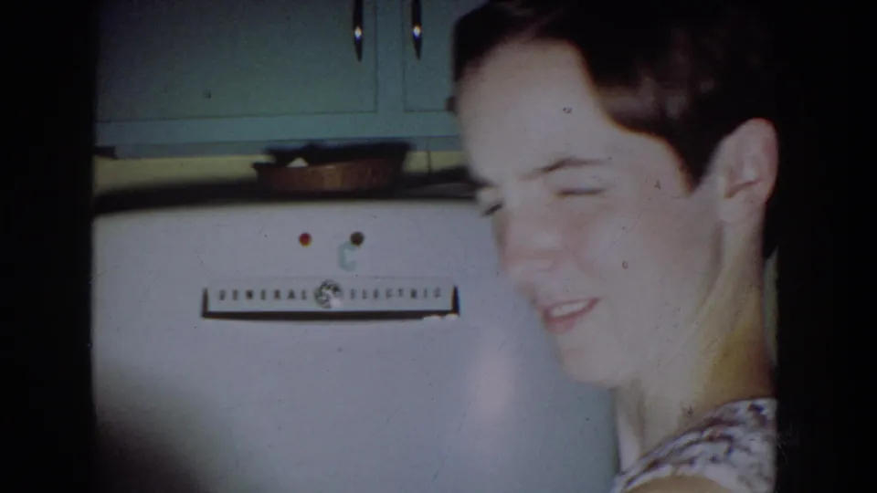 1965 kitchen frame showing a seated young man near a white General Electric refrigerator and upper cabinets.