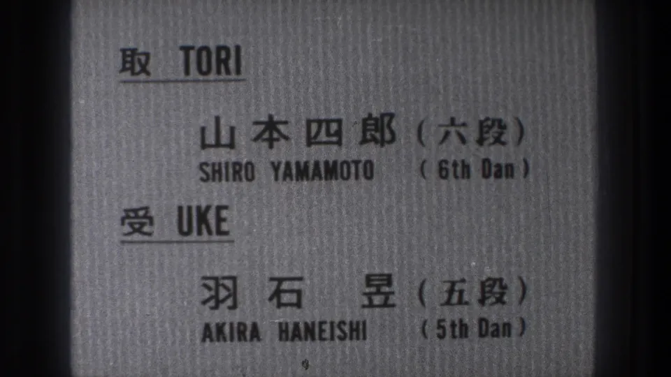 Close 8mm frame of the bilingual martial arts card, with names and dan ranks printed in black.