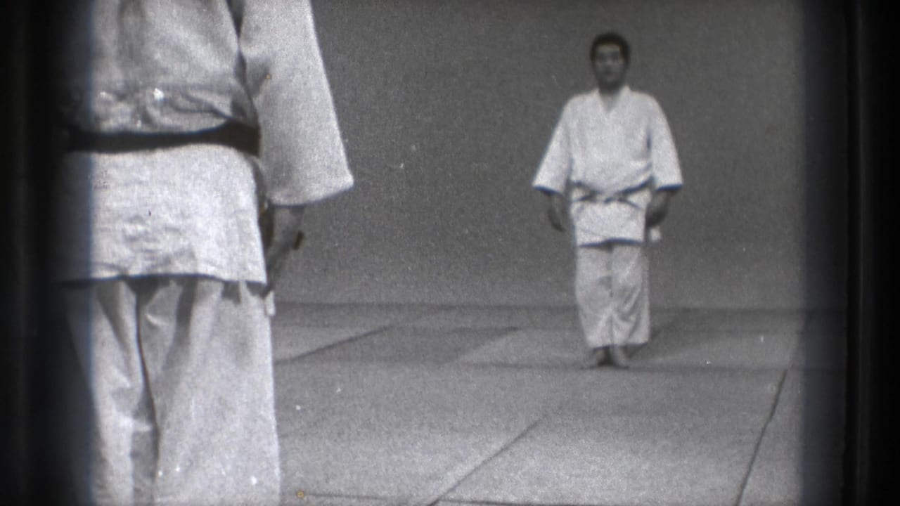 1971 Tokyo home-movie frame showing a martial arts instructor in a white gi and dark belt facing the camera on dojo mats.