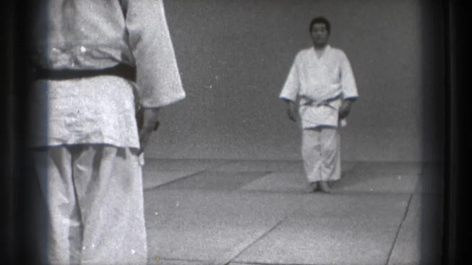 1971 Tokyo home-movie frame showing a martial arts instructor in a white gi and dark belt facing the camera on dojo mats.
