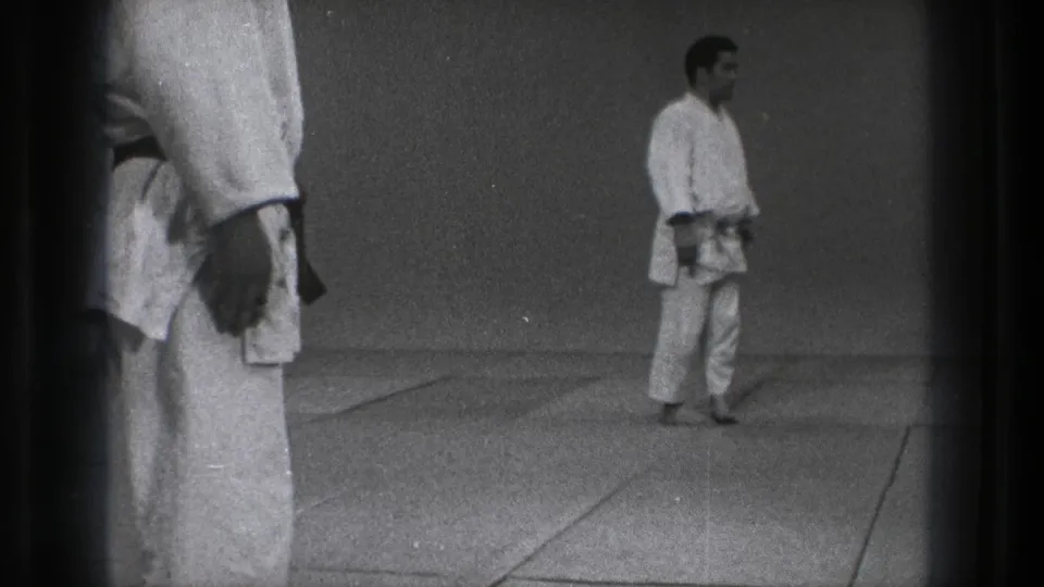 1971 Tokyo frame with the instructor turned to the side in a white gi, seen across a simple matted training room.