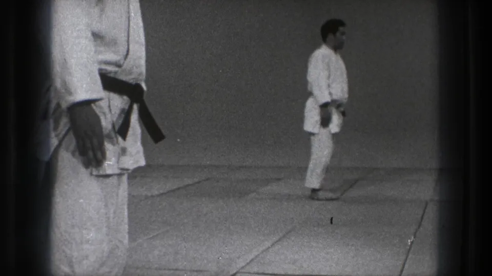 Side view of a martial arts instructor in a white gi and dark belt on a matted floor, with 8mm frame edges visible.