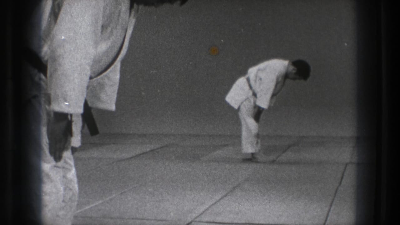 In 1971 Tokyo footage, a man in a white martial arts gi bows on a mat while a second black-belt figure stands partly cropped nearby.
