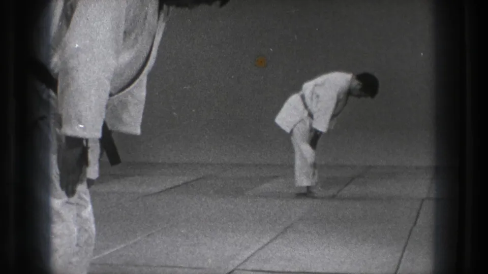 In 1971 Tokyo footage, a man in a white martial arts gi bows on a mat while a second black-belt figure stands partly cropped nearby.