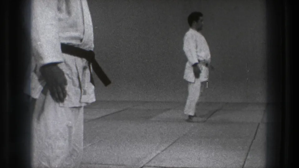 A black-belt martial arts practitioner stands in profile on a dojo mat in the supplied 1971 Tokyo 8mm footage.