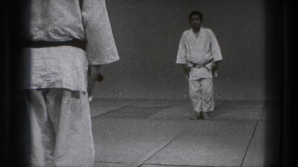 The practitioner stands facing the camera in a sparse dojo-like room, with an observing figure partly cropped in the foreground.