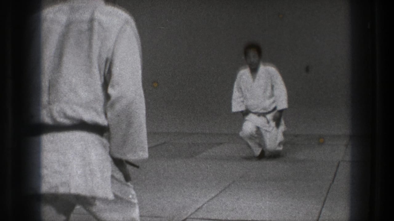 Two men in white gi on dojo mats, one standing in the foreground and one kneeling ahead.
