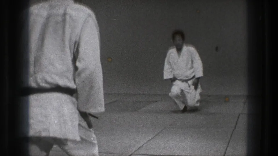 1971 Tokyo 8mm frame showing one man in a gi from behind while another kneels on the dojo mat facing him.