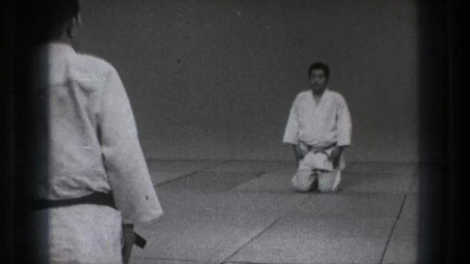1971 Tokyo 8mm frame with a foreground practitioner seen from behind and a second man kneeling formally on mats.