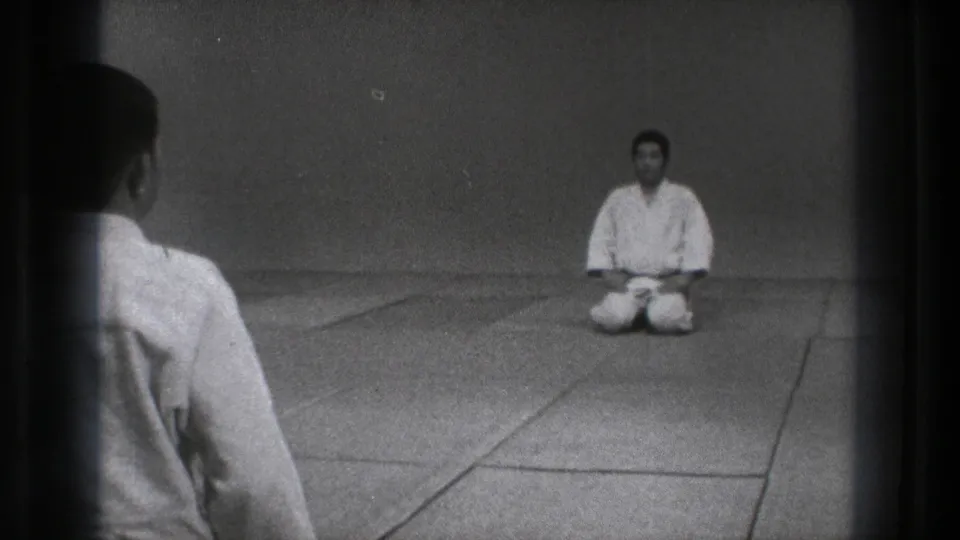 1971 Tokyo 8mm frame showing a foreground practitioner and a kneeling man facing each other in a spare dojo.