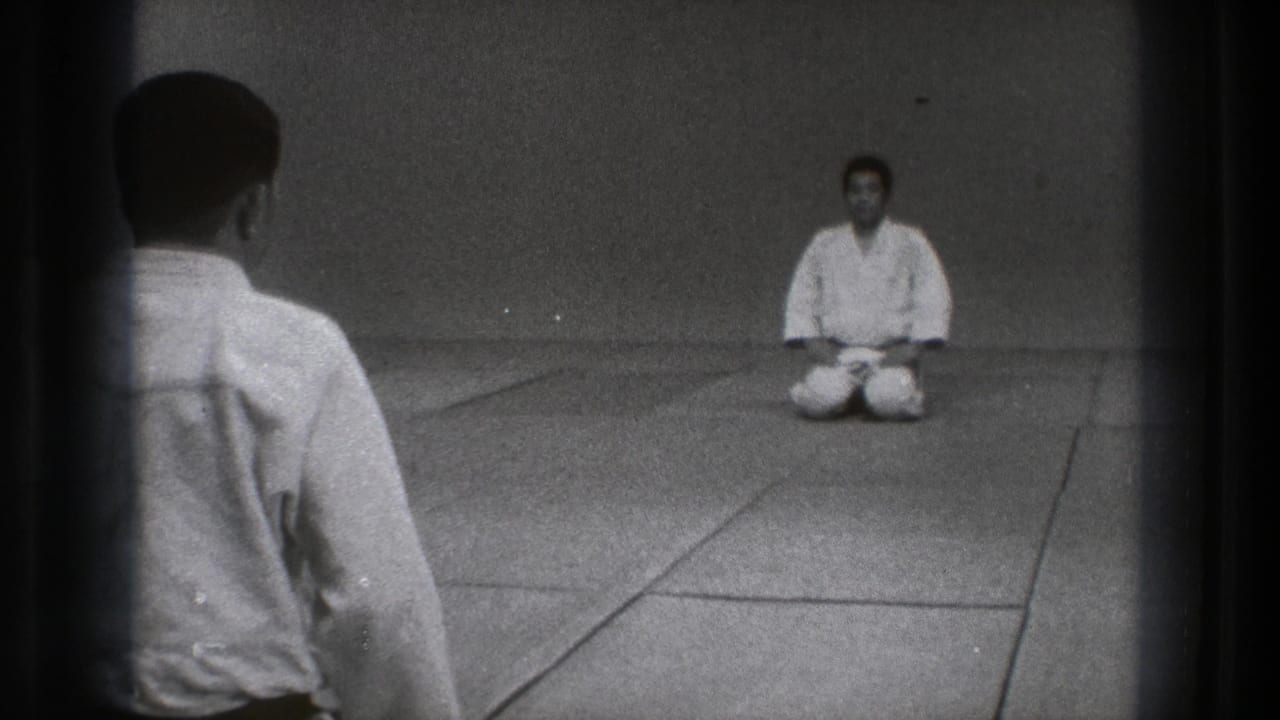 Two men in white gi face each other on tatami mats, one seen from behind and one seated in seiza.