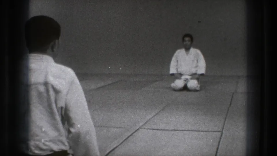 1971 Tokyo 8mm frame showing two martial arts practitioners in white gi facing each other across a plain dojo floor.