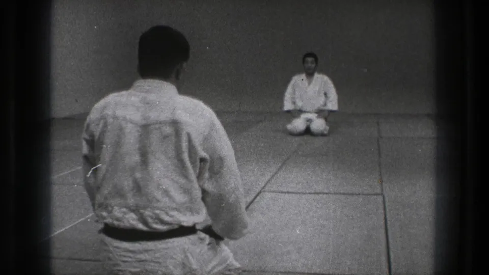1971 Tokyo home-movie frame of two martial arts practitioners facing each other in seiza inside a plain dojo.
