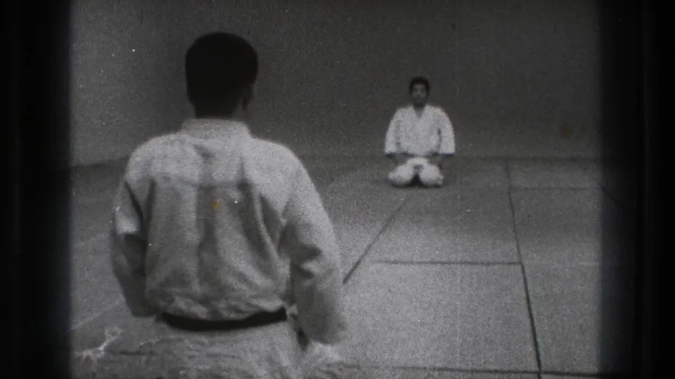 1971 Tokyo 8mm frame showing two men in karate uniforms kneeling across a plain dojo mat.