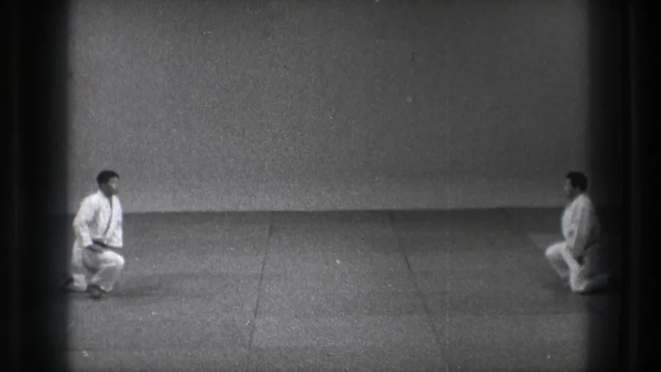 1971 Tokyo dojo frame showing two men in white gi shifting from kneeling toward standing.