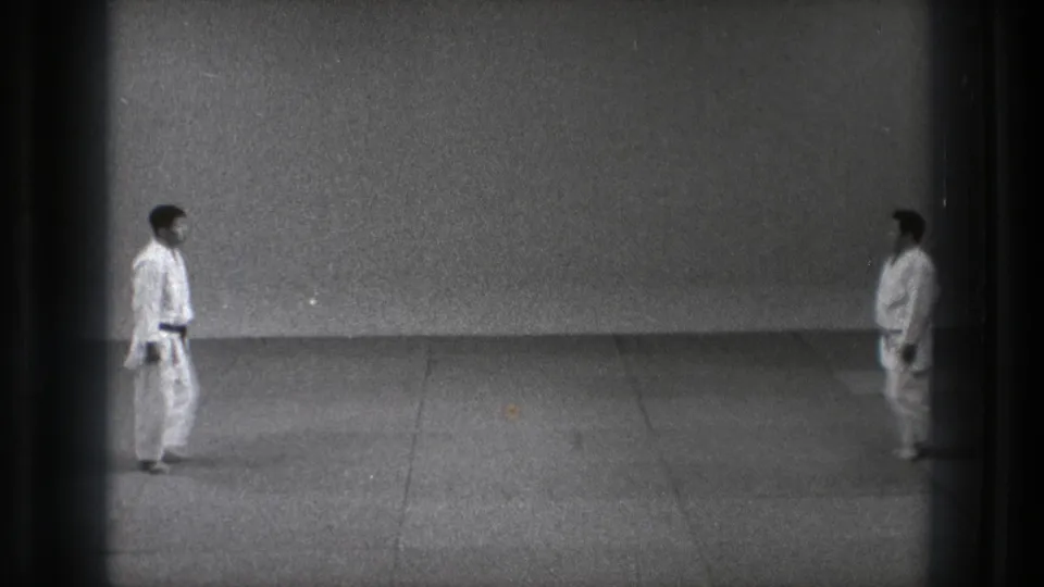In a Tokyo dojo setting from the 1971 clip, two adult men in white gi and dark belts stand apart on a plain mat.