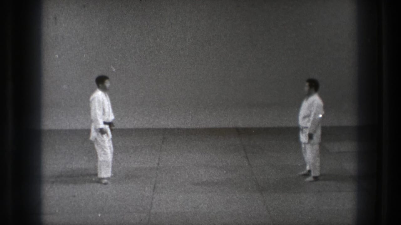 Two men in gi stand facing each other in a simple indoor training space.