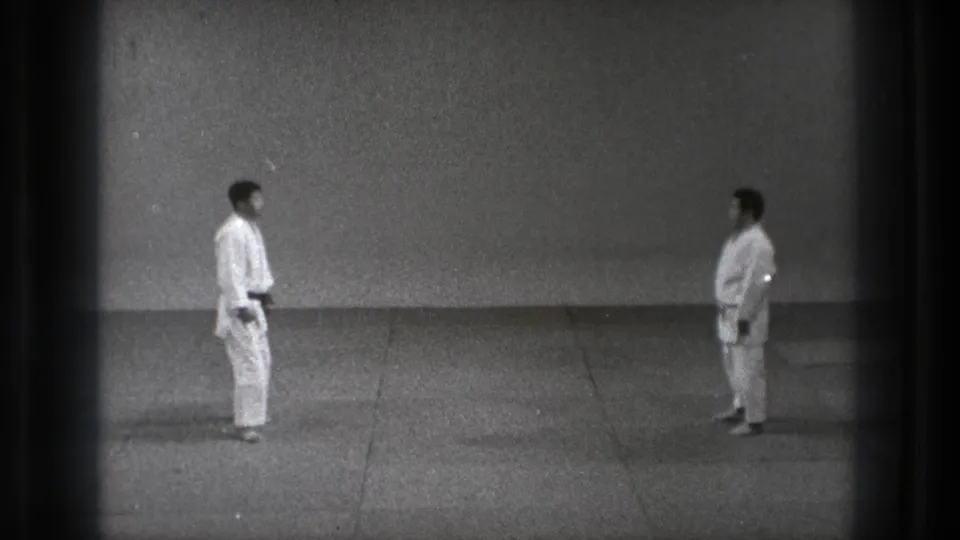 Two adult martial arts practitioners in white gi and dark belts face one another on a bare mat in the Tokyo 1971 footage.