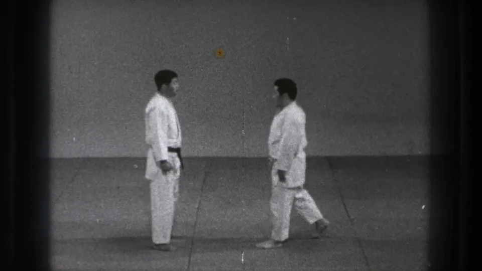 In Tokyo 1971 footage, two men in white gi and dark belts stand facing each other on a plain training mat.