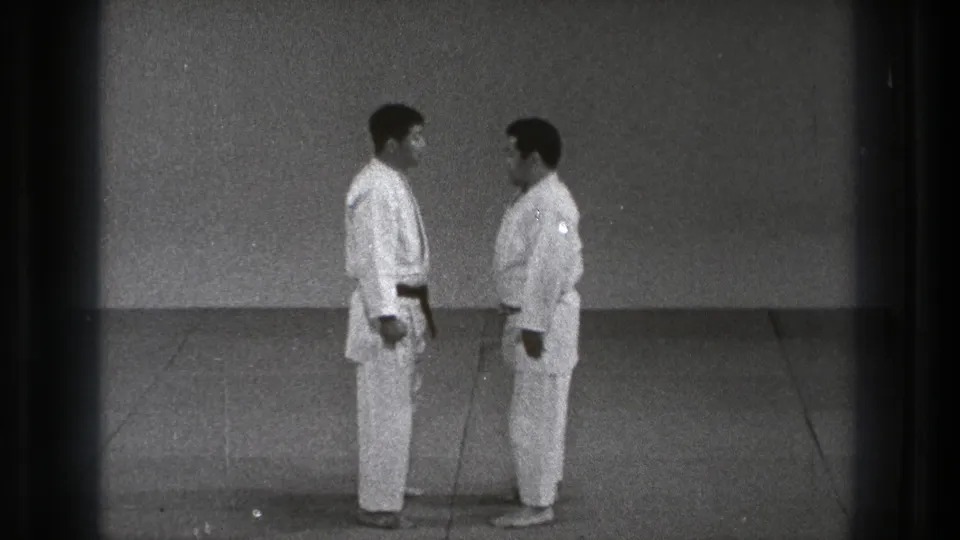 Two adult martial arts practitioners in white gi stand close together on a sparse mat in the Tokyo 1971 clip.