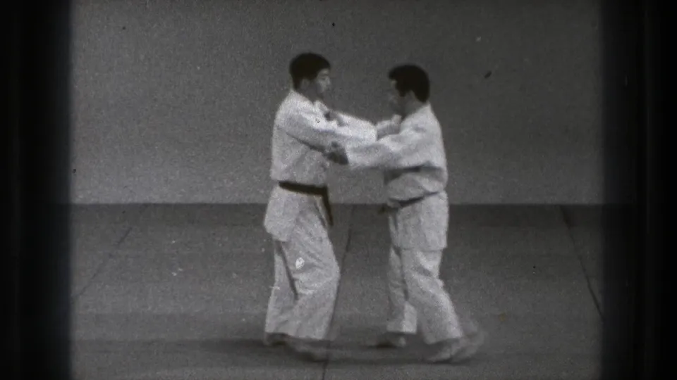 Two men in white gi and dark belts demonstrate a controlled hand technique on a mat in the 1971 Tokyo footage.