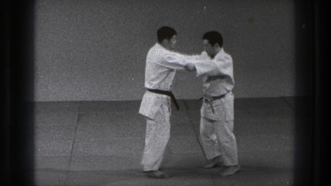 In supported 1971 Tokyo footage, two adult men in white gi face each other in a judo grip on an indoor mat.