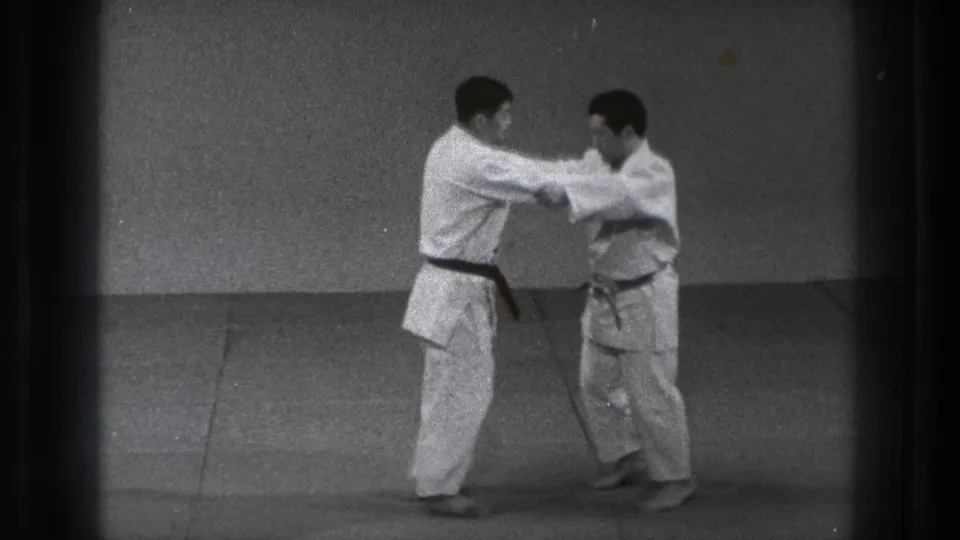 In supported 1971 Tokyo footage, two adult men in white gi face each other in a judo grip on an indoor mat.