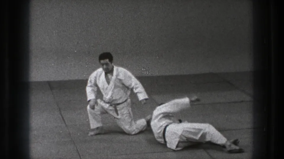 After the throw, one man kneels on the padded floor while his partner lies on his back in the indoor training space.