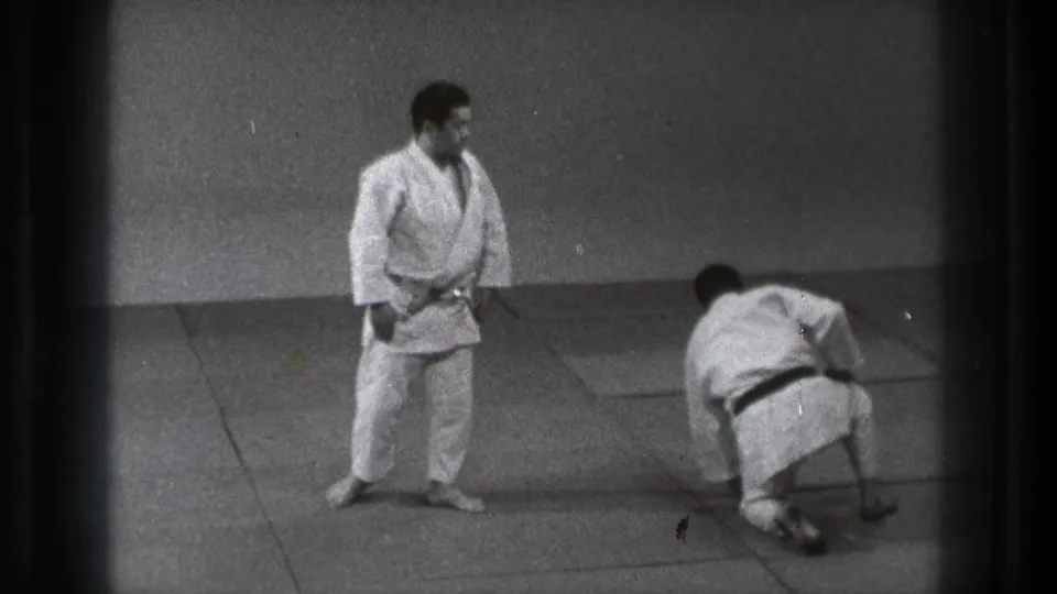 One practitioner stands and watches as his partner pushes up from the mat during the reset after a judo throw.