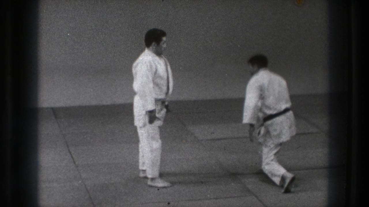 Two men in white judo uniforms stand on indoor mats as one bends forward.