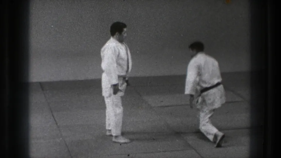 In 1971 Tokyo footage, two adult martial artists in white gi stand on dojo mats while one bends near his partner.
