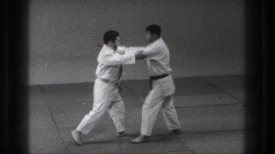 Two adult martial artists in white gi grip shoulders and sleeves during a controlled judo practice in 1971 Tokyo.
