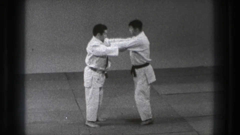 In Tokyo 1971 footage, two men in white gi stand on a mat and grip each other during martial arts practice.