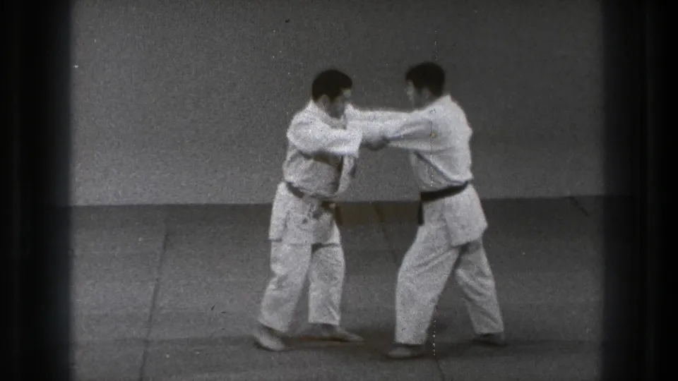 Two martial arts practitioners in white gi maintain arm contact while shifting stance on an indoor mat in Tokyo 1971 footage.