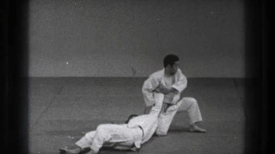 A gi-clad practitioner drops to one knee while the other is thrown or lowered onto the mat during the technique.