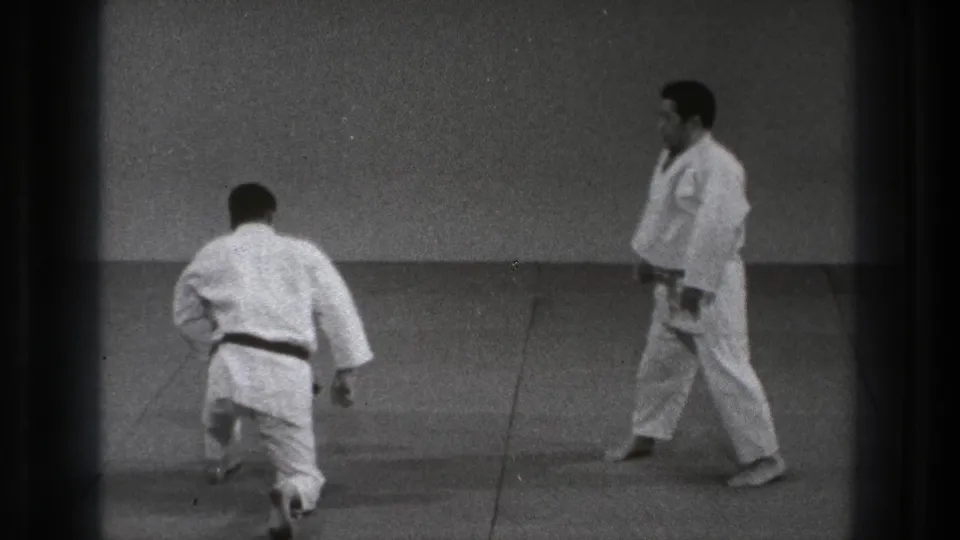 Two adult men in white gi practice across a matted floor in the Tokyo 1971 martial arts sequence.