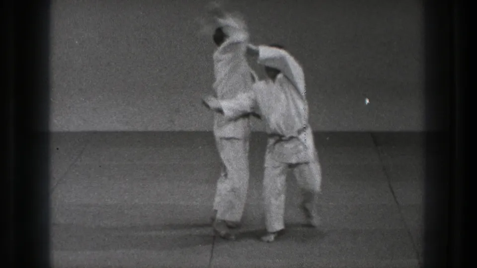In Tokyo 1971 footage, two adult men in white gi stand close as one begins a martial arts throwing technique on a plain mat.