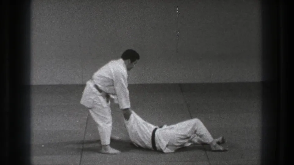The throw demonstration continues as one man stands over his partner, who lies on the mat with a dark belt visible.