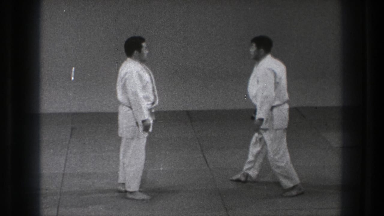 Two men in gi face each other on an indoor mat.