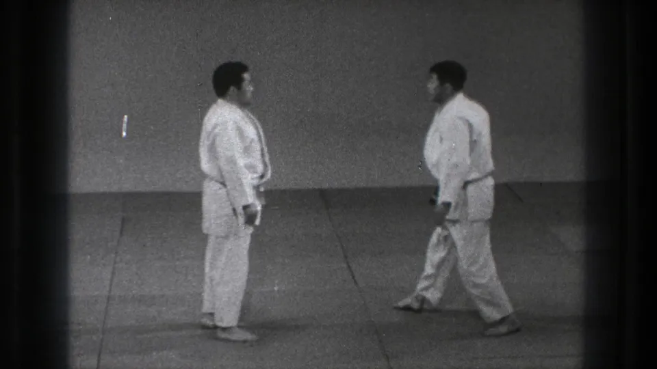 Two adult men in white gi face one another in a sparse indoor training space in the supplied Tokyo, 1971 home-movie clip.