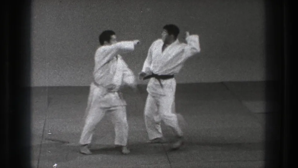 A practitioner in a white gi extends a strike as the other turns or reacts during indoor karate practice in the supplied 1971 Tokyo footage.