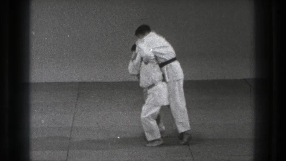 In 1971 Tokyo footage, two adult men in white martial arts uniforms grip each other during a dojo throw drill.