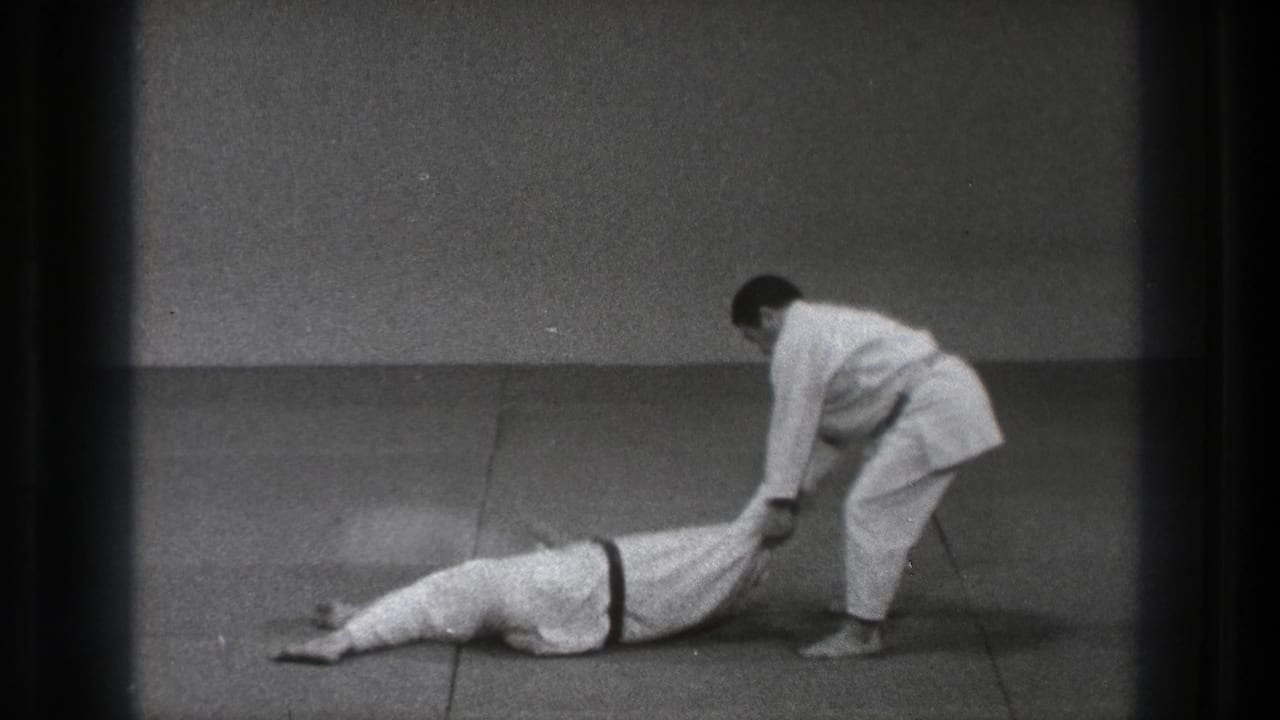 One man lies face down while another holds his legs on the mat.