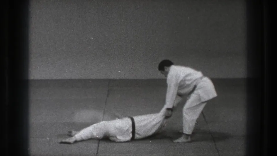 A martial arts practitioner in a white gi holds his partner's legs after a throw in the Tokyo training room.