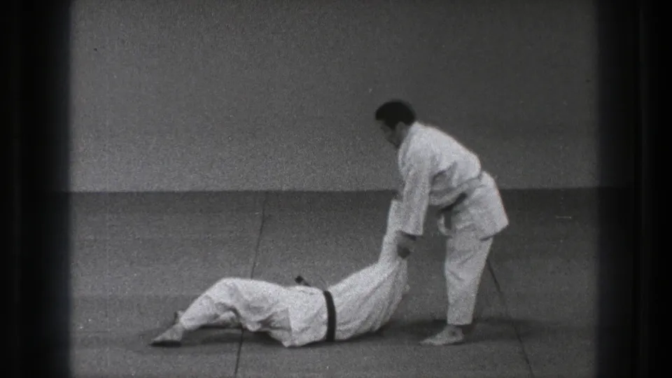 The throw sequence continues as one man in a gi holds the legs of his partner lying across the mat.