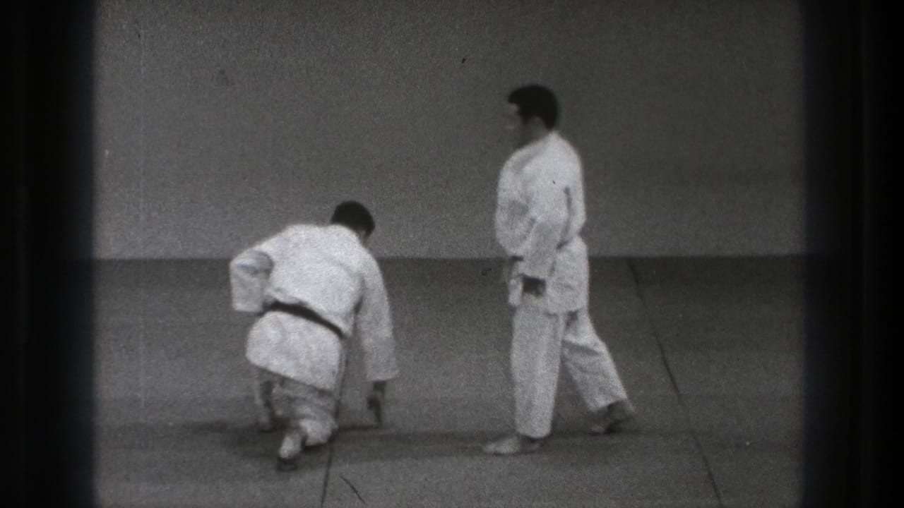Two men in karate gi on mats, one crouching low while the other stands nearby.