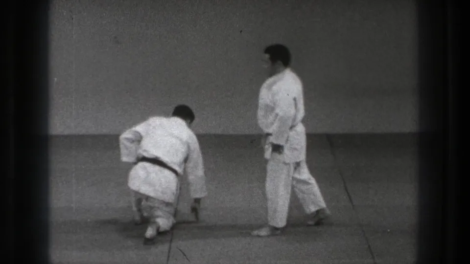 1971 Tokyo 8mm frame of two adult men in white karate gi on floor mats, with one crouched low and the other standing to the side.